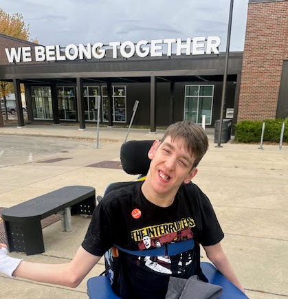 Justin smiling, seated in wheelchair, I voted sticker on shirt,, building with We Belong Together sign behind him