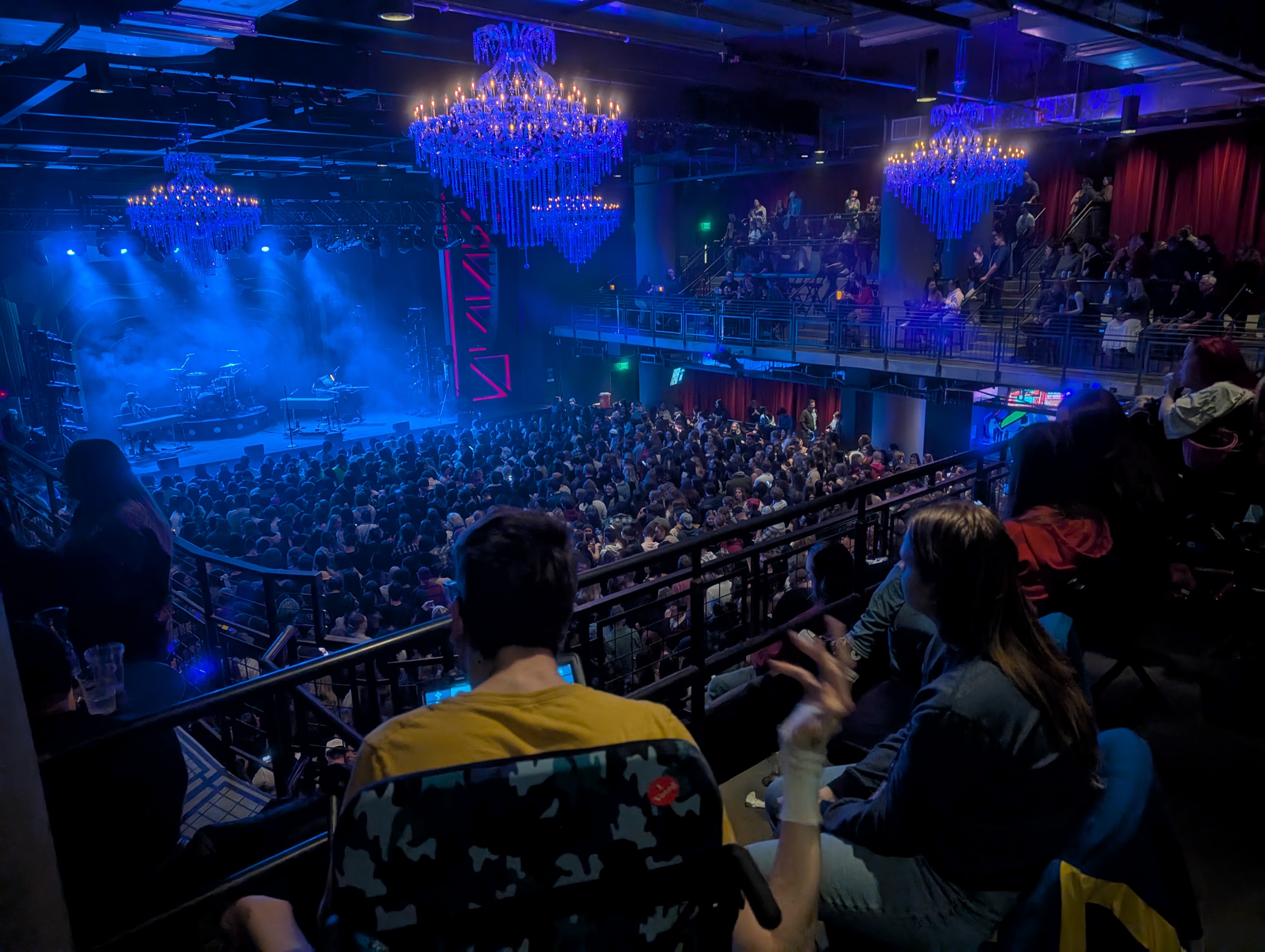 Back of man in wheelchair in balcony looking towards stage, crowd on floor