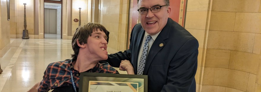 Justin smiling at Senator Hoffman who is holding Justin's Youth Award certificate and smiling at the camera