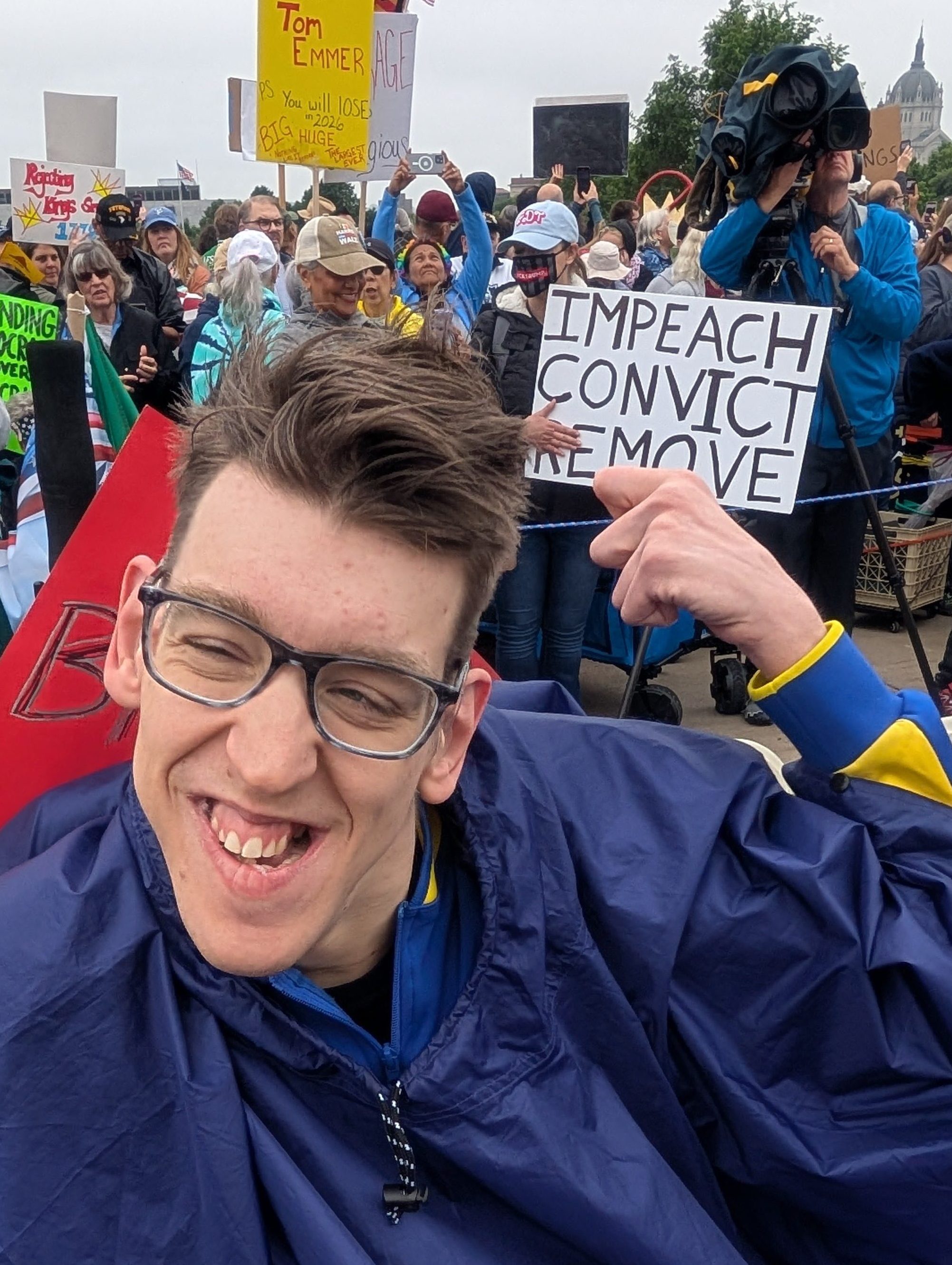 Justin smiling wearing blue poncho, at No Kings rally with crowd behind him