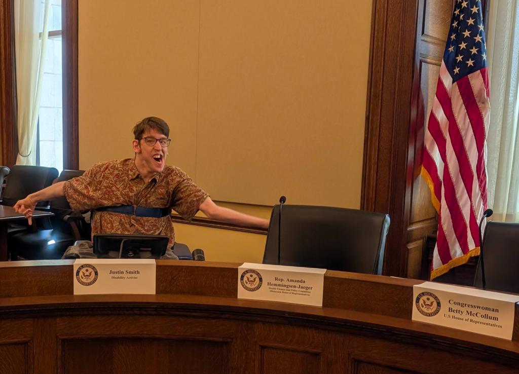 Justin, a young man in wheelchair smiling with a look that says "IReady to go!", name tags and seats around a Senate hearing table, American flag