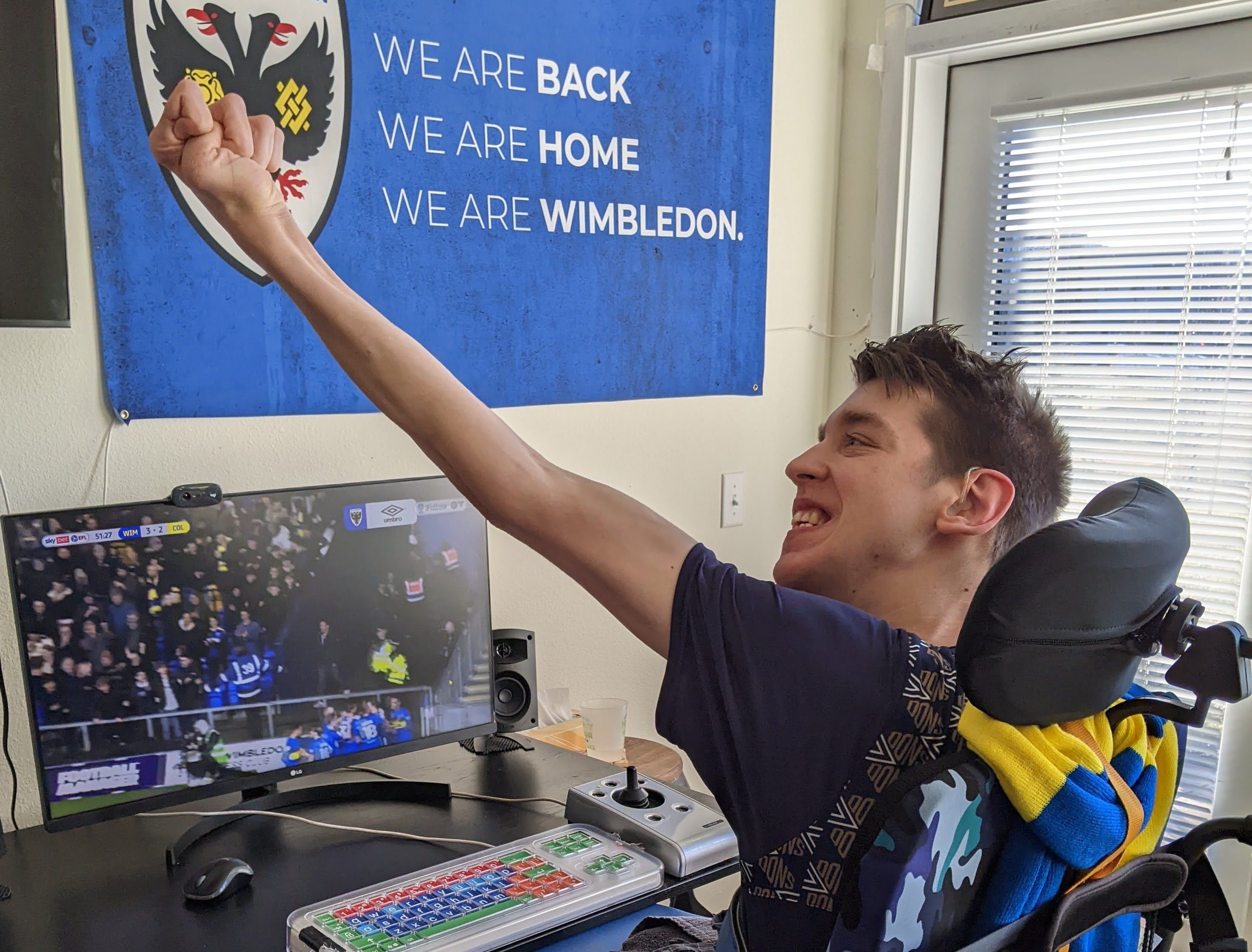 Justin smiling at soccer match on computer monitor with arm raised in the air cheering a goal. AFC Wimbledon flag on wall says "we are back, we are home, we are Wimbledon"