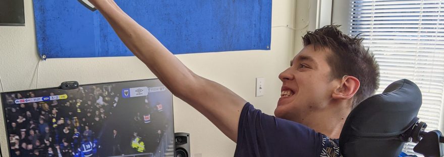 Justin smiling at soccer match on computer monitor with arm raised in the air cheering a goal. AFC Wimbledon flag on wall says "we are back, we are home, we are Wimbledon"