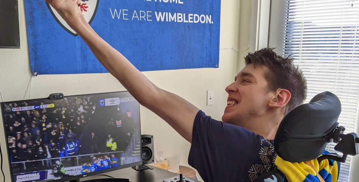 Justin smiling at soccer match on computer monitor with arm raised in the air cheering a goal. AFC Wimbledon flag on wall says "we are back, we are home, we are Wimbledon"