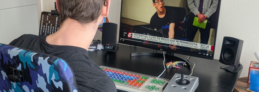 Justin sitting at his computer with accessible keyboard watching a kstp news story that's showing a picture of him in his wheelchair