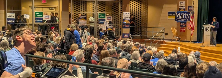 Peggy Flanagan speaking on stage at podium, Justin in a wheelchair smiling in the back of the auditorium filled with people, political signs posted on walls