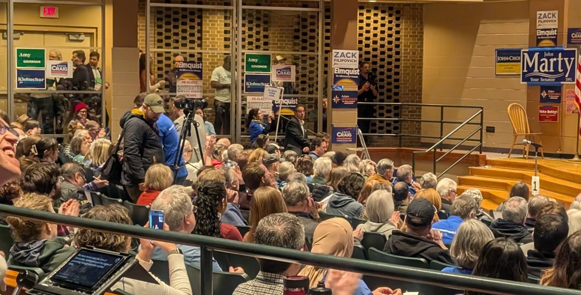 Peggy Flanagan speaking on stage at podium, Justin in a wheelchair smiling in the back of the auditorium filled with people, political signs posted on walls