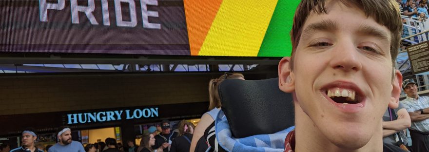Justin smiling with MN Loons scarf around his neck, signage behind says Pride with rainbow colors, people standing behind and in bleachers