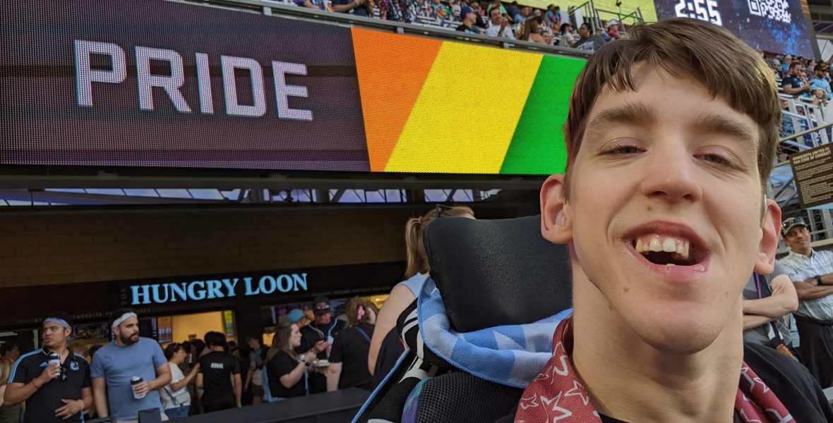 Justin smiling with MN Loons scarf around his neck, signage behind says Pride with rainbow colors, people standing behind and in bleachers