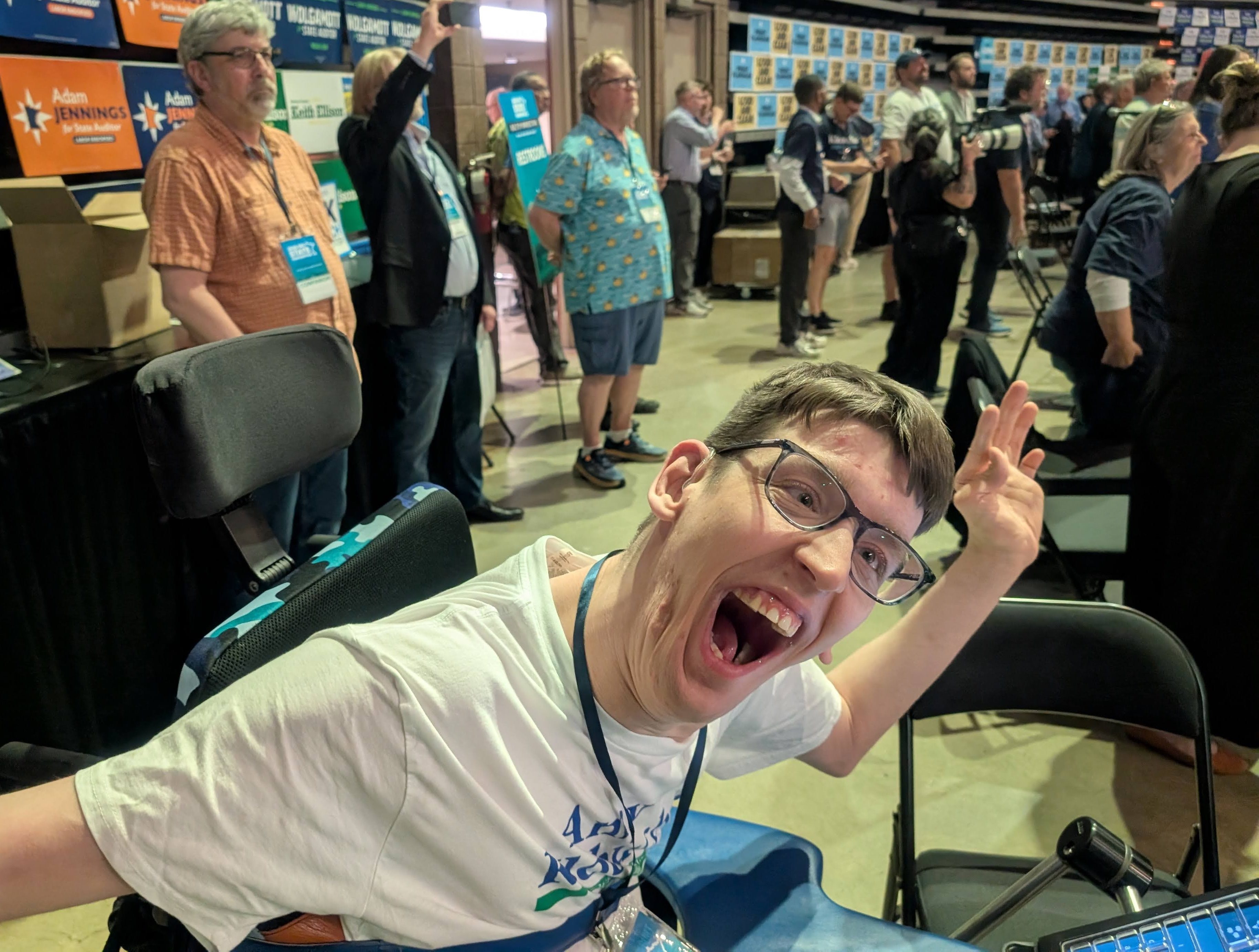 Justin, a young man smiling in excitement, seated in power wheelchair at table and wearing Amy Klobuchar shirt, people standing behind him, convention signs posted on wall