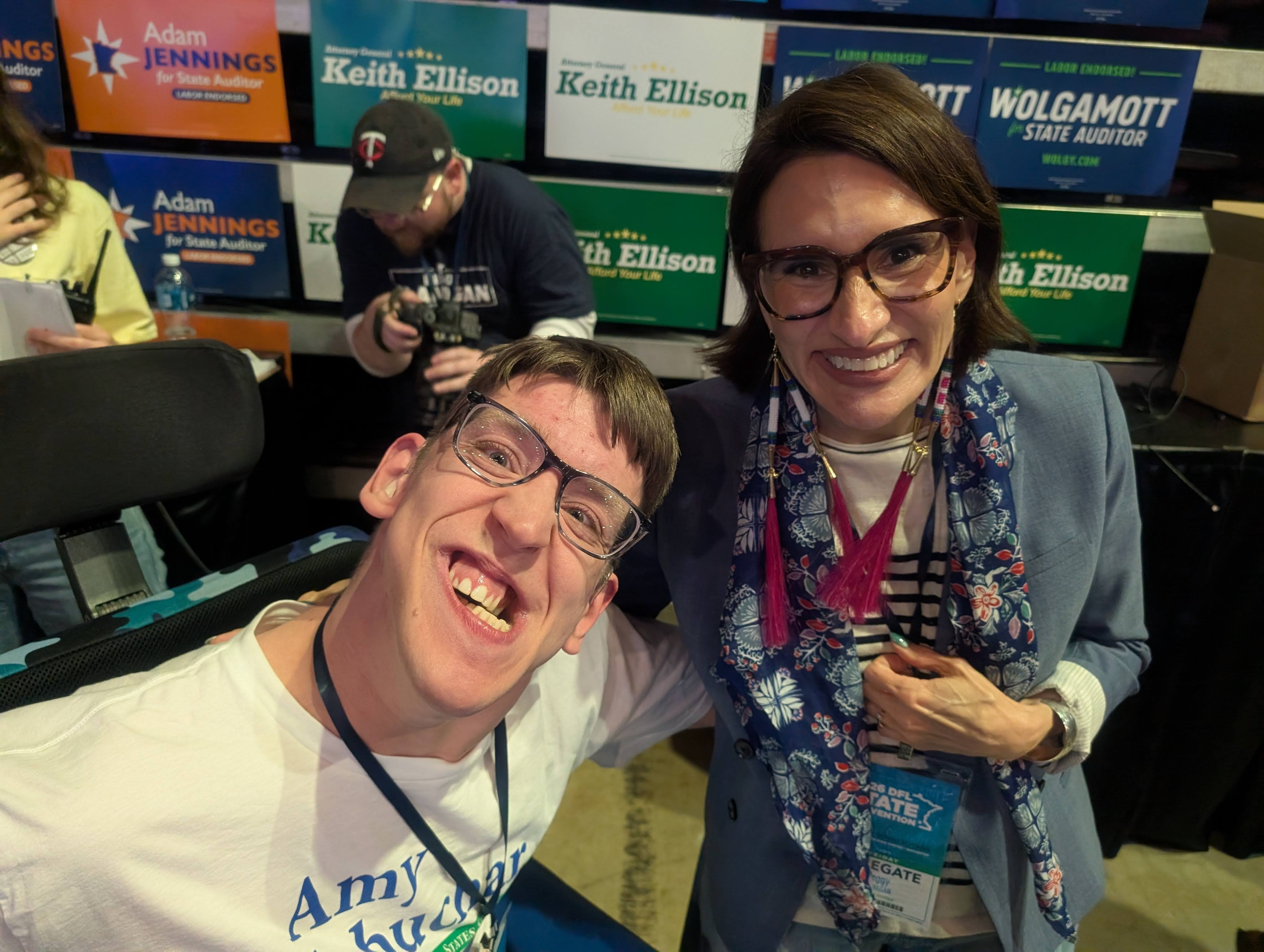 Justin, young man smiling and seated in power wheelchair, Peggy Flanagan leaning in and smiling next to him, convention posters for various democratic candidates behind them