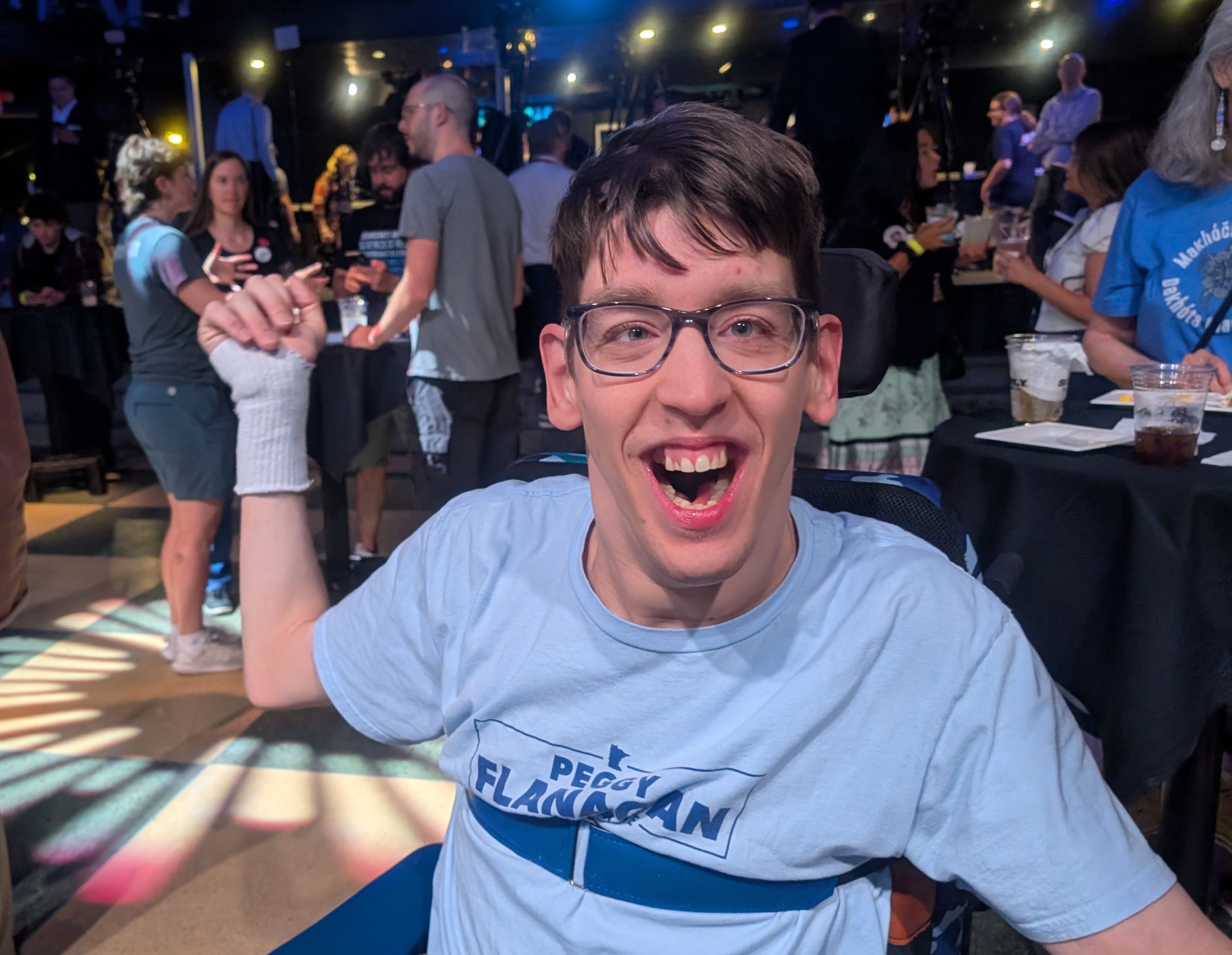 Young man smiling seated in wheelchair and wearing Peggy Flanagan shirt, groups of people behind