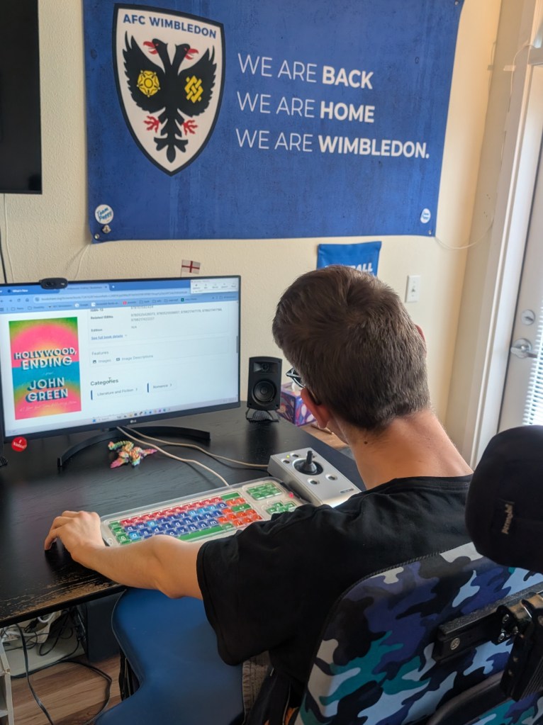Back of young man in wheelchair looking at Hollywood, Ending book image on Bookshare.org on computer monitor, adapted keyboard and joystick mouse on table, AFC Wimbledon flag on wall says we are back, we are home, we are wimbledon