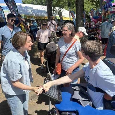 Senator Amy Klobuchar shaking hands with Justin who is a young man seated in a power wheelchair, he's looking at his mom who is greeting Amy, state fair crowd in background