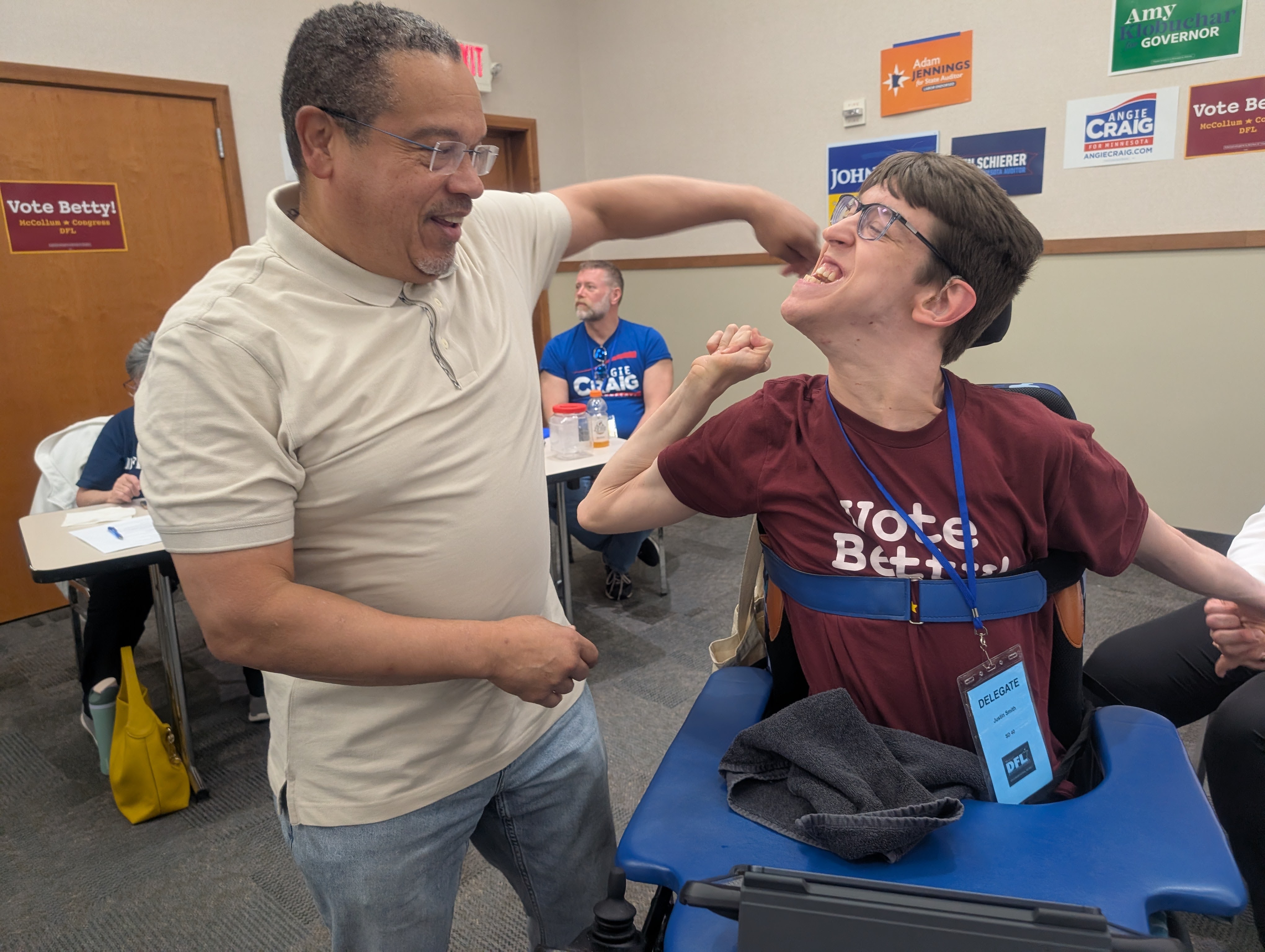 Attorney General Keith Ellison reaching towards Justin who is seated in wheelchair wearing a vote betty shirt, candidate signs hanging on wall behind them